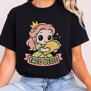 May include: Black t-shirt with a cartoon princess wearing a crown and holding a taco. The text "TACO BELLE" is on a pink banner. The princess has pink hair and is wearing a green dress.