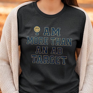 May include: Dark gray t-shirt with the text "I AM MORE THAN AN AD TARGET" in blue and gold lettering. A yellow smiley face graphic is above the text. The shirt is a crew neck style.