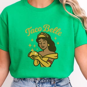 May include: Green t-shirt with the text "Taco Belle" in gold script above a cartoon illustration. The illustration depicts a woman in a yellow dress holding a taco, with a taco-shaped crown and star accents.