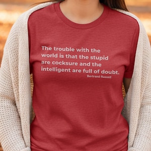 May include: A red t-shirt featuring a quote by Bertrand Russell: "The trouble with the world is that the stupid are cocksure and the intelligent are full of doubt." The text is in white. A cream cardigan is worn.