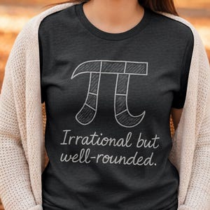 Puede incluir: Camiseta gris oscuro con un boceto blanco del símbolo Pi. Debajo, el texto dice "Irrational but well-rounded". La camiseta es de cuello redondo.