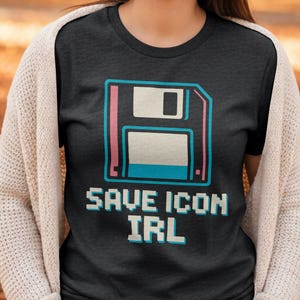 Puede incluir: Camiseta gris oscuro con un diseño pixelado de disquete con detalles en rosa y azul. El texto "SAVE ICON IRL" se muestra debajo en una fuente pixelada. Es una prenda de vestir informal.