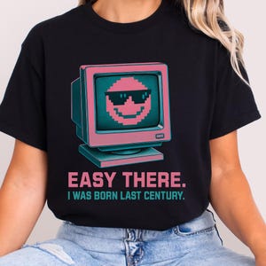May include: Black t-shirt with a retro computer graphic. The graphic shows a pixelated smiley face wearing sunglasses. Below the graphic is the text "EASY THERE. I WAS BORN LAST CENTURY." The design has a vintage aesthetic, reminiscent of the 1980s or 1990s.