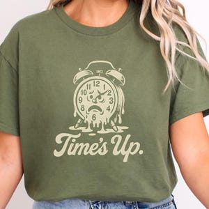 May include: Olive green t-shirt featuring a distressed graphic of a melting alarm clock with the words "Time's Up" in a vintage font. The clock face shows the time as approximately 1:00. The shirt is a casual wear item.