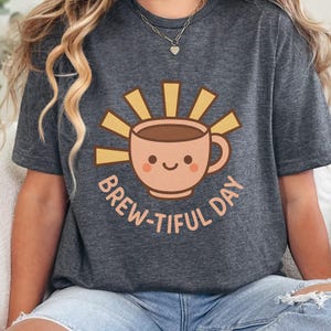 Peut inclure: T-shirt gris foncé avec une tasse de café souriante et un motif de soleil. Le texte "BREW-TIFUL DAY" est imprimé autour de la tasse. Le t-shirt est fait d'une matière douce. Un haut décontracté pour les amateurs de café.