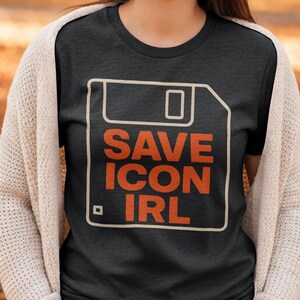 May include: Dark grey t-shirt with a retro floppy disk graphic and the orange text "SAVE ICON IRL". The design is outlined in white. The t-shirt is a classic crew neck style.