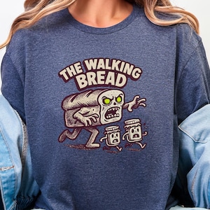 Puede incluir: Sudadera azul marino con un diseño de dibujos animados. El gráfico muestra un zombie de pan con ojos verdes y el texto "THE WALKING BREAD". Dos tarros de mermelada huyen. El diseño utiliza colores marrón, blanco y negro.