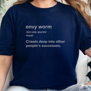 以下が含まれることがあります： 「envy worm」と定義が書かれたネイビーブルーのTシャツ。定義は「他人の成功に深く入り込む」です。カジュアルなトップスです。