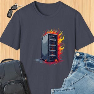 Puede incluir: Una camiseta azul marino con una ilustración de dibujos animados de un servidor en llamas. El servidor es de color gris oscuro con luces rojas y está rodeado de llamas naranjas.