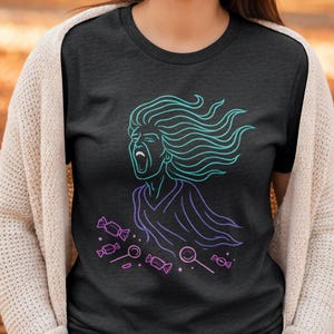 Puede incluir: Camiseta gris oscuro con una ilustración de neón de una figura gritando con el pelo suelto. El diseño incluye caramelos y piruletas rosas y moradas. La camiseta es de cuello redondo y la lleva una persona.
