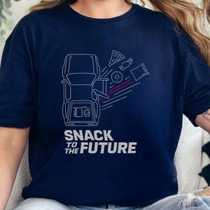 Op de afbeelding: Marineblauw T-shirt met een witte afbeelding van een auto en snacks, waaronder pizza, snoep, een donut en een zak chips. De tekst "SNACK TO THE FUTURE" staat eronder.