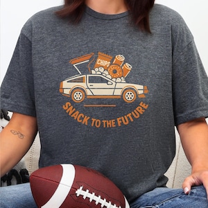 Op de afbeelding: Donkergrijs t-shirt met een afbeelding van een auto vol snacks, waaronder chips en donuts. De oranje tekst luidt "SNACK TO THE FUTURE". Het t-shirt heeft een casual stijl.