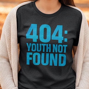 Könnte beinhalten: Dunkelgraues T-Shirt mit dem Text "404: YOUTH NOT FOUND" in leuchtend blauen Blockbuchstaben. Die Person trägt einen cremefarbenen Cardigan. Der Hintergrund ist verschwommen mit Herbstfarben.