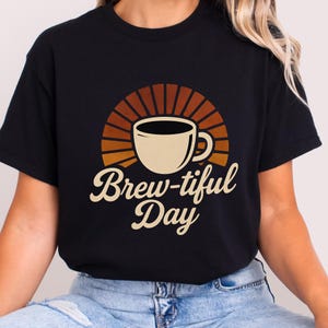 Peut inclure: T-shirt noir avec un motif de tasse à café et un fond en forme d'éclat de soleil marron et orange. Le texte "Brew-tiful Day" est écrit en cursive en dessous de la tasse. Le t-shirt est en matière souple.