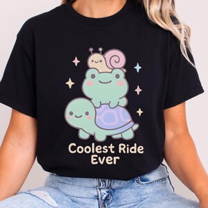 Puede incluir: Camiseta negra con un diseño de dibujos animados de una tortuga, una rana y un caracol apilados. La rana está sobre la espalda de la tortuga, y el caracol está sobre la cabeza de la rana. El texto "Coolest Ride Ever" está debajo.