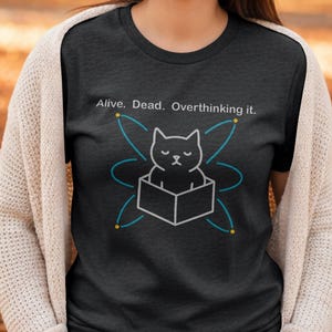 Puede incluir: Camiseta gris oscuro con el texto "Alive. Dead. Overthinking it." y un gráfico de un gato en una caja, rodeado por un diseño similar a un átomo. El gato es blanco y negro, y el diseño tiene detalles en azul y amarillo.