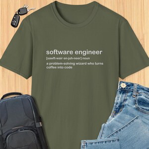 Könnte beinhalten: Olivgrünes T-Shirt mit der weißen Definition von "Softwareentwickler". Die Definition enthält die phonetische Aussprache und den Satz "ein Problemlösungszauberer, der Kaffee in Code verwandelt."