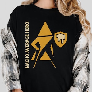 Puede incluir: Camiseta negra con un gráfico dorado de una figura de superhéroe y el texto "NACHO AVERAGE HERO" en dorado. La figura tiene una cabeza triangular, un escudo y una capa. La camiseta se lleva sobre una camisa a cuadros.