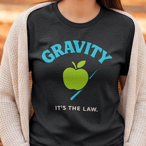 May include: Dark gray t-shirt with the word "GRAVITY" in light blue above a green apple graphic. Below the apple, the text reads "IT'S THE LAW." The shirt is a casual style, suitable for everyday wear.