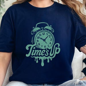 Puede incluir: Camiseta azul marino con un gráfico de despertador verde menta y el texto "Time's Up". El diseño del reloj incluye los números del 1 al 12. La camiseta está hecha de un material suave.