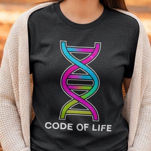 Op de afbeelding: Donkergrijs T-shirt met een kleurrijk DNA-dubbele helix ontwerp. De afbeelding bevat blauwe, roze en groene tinten. De woorden "CODE OF LIFE" staan in het wit onder de helix. Het shirt heeft een ronde hals.