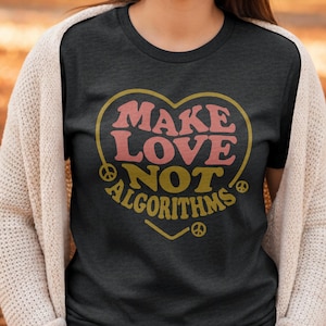 May include: Dark gray t-shirt with a heart-shaped design. The design features the words "MAKE LOVE NOT ALGORITHMS" in pink and yellow, with peace signs. The shirt is worn with a cream-colored cardigan.