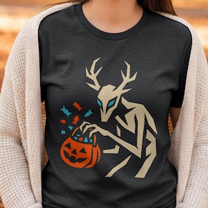 May include: Dark gray t-shirt featuring a stylized figure with antlers and blue eyes, holding an orange jack-o'-lantern bucket. The design includes falling candy in blue, orange, and red. A Halloween-themed graphic tee.