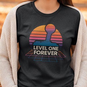 Könnte beinhalten: Dunkelgraues T-Shirt mit einem Retro-Design, das einen Joystick vor einem Sonnenuntergang in Orange, Pink und Lila zeigt. Der Text "LEVEL ONE FOREVER" ist darunter in Weiß dargestellt.