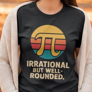 Puede incluir: Camiseta gris oscuro con un gráfico del símbolo Pi en un círculo con un diseño retro de puesta de sol. El texto "IRRATIONAL BUT WELL-ROUNDED" está debajo. El diseño tiene colores amarillo, naranja, rojo y azul.