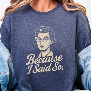 Könnte beinhalten: Marineblaues T-Shirt mit einer Retro-Illustration einer Frau mit Cat-Eye-Brille. Der Text "Because I Said So" steht unter der Illustration in einer Schreibschrift. Das T-Shirt ist aus einem weichen Material.