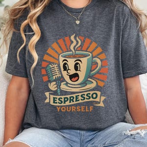 Peut inclure: T-shirt gris foncé avec un dessin de tasse à café de dessin animé tenant un microphone, avec de la vapeur et un visage souriant. Le design comprend un soleil rétro et les mots "Espresso Yourself" sur une bannière.