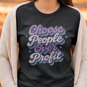 May include: Dark gray t-shirt with the text "Choose People Over Profit" in a retro font. The text is in white, purple, and blue, with star accents. The shirt is a crew neck style.