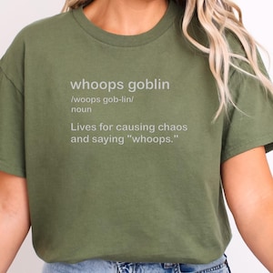 Puede incluir: Camiseta verde oliva con la frase "whoops goblin" en gris claro. La definición incluye la pronunciación, la palabra "sustantivo" y la frase "Vive para causar caos y decir 'whoops.'"