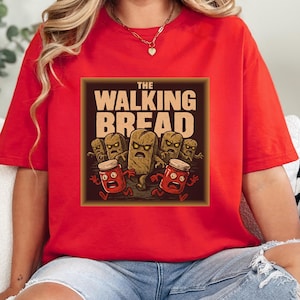 Puede incluir: Camiseta roja con un gráfico humorístico. El diseño presenta el texto "THE WALKING BREAD" con personajes de pan de dibujos animados y botes de mermelada corriendo. La camiseta es de color rojo brillante.