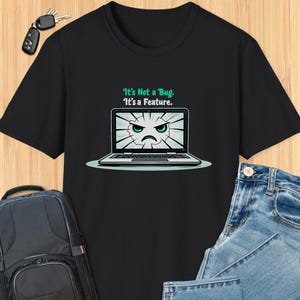 以下が含まれることがあります： 白いラップトップのグラフィックがプリントされた黒いTシャツ。ラップトップの画面には、怒った表情の漫画の顔が描かれています。シャツのテキストは「It's Not a Bug. It's a Feature.」です。
