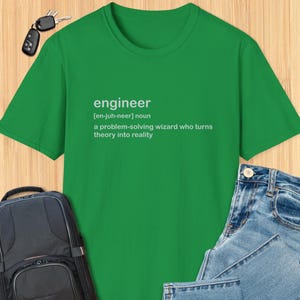 以下が含まれることがあります： 「engineer」の文字が白でプリントされた緑色のTシャツ。発音と定義も記載されています。「問題を解決し、理論を現実にする魔法使い」という意味です。青いジーンズ、バックパック、鍵も写っています。
