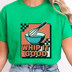 Puede incluir: Camiseta verde con un diseño gráfico retro. El diseño incluye un cuenco turquesa, un batidor y el texto "WHIP IT GOOD" sobre un fondo melocotón, con acentos a cuadros blancos y negros. Un tema divertido relacionado con la comida.