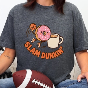 May include: Dark gray t-shirt with a playful graphic of a donut dribbling a basketball, a coffee mug, and the text "SLAM DUNKIN'". A football is visible in the foreground. The shirt is made of a soft material.