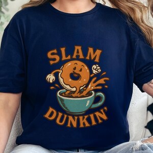 May include: Navy blue t-shirt featuring a cartoon donut dunking into a teal coffee cup. The words "SLAM DUNKIN'" are in orange above and below the donut illustration. The donut has a smiling face and is splashing into the coffee.