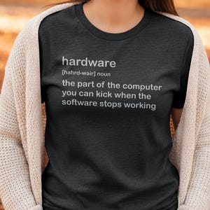 May include: Dark grey t-shirt with the definition of "hardware" printed in white. The definition reads: "the part of the computer you can kick when the software stops working." The shirt is worn by a person wearing a cream-coloured cardigan.