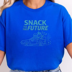 以下が含まれることがあります： ロイヤルブルーのTシャツに、ティールグリーンのグラフィック。グラフィックは、ファストフードを運ぶ車と「SNACK TO THE FUTURE」の文字が特徴です。デザインには、ハンバーガー、フライドポテト、アイスクリームが含まれています。