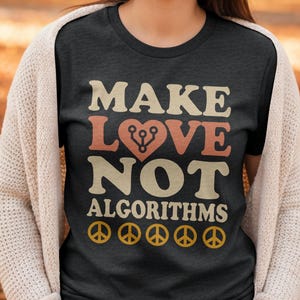 Op de afbeelding: Donkergrijs t-shirt met de tekst "MAKE LOVE NOT ALGORITHMS" in beige en koraalkleurige letters. Het woord "LOVE" heeft een hart met een printplaatontwerp. Daaronder staan vijf gele vredestekens.