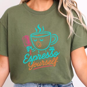 Peut inclure: T-shirt vert olive avec une tasse à café de dessin animé chantant dans un microphone rose. L'expression "Espresso Yourself" est écrite en script bleu sarcelle et orange en dessous de la tasse. Le motif est centré sur le t-shirt.
