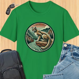Pode incluir: Camiseta verde com um gráfico de um leão da montanha sentado em uma rocha em um cenário de floresta. O gráfico está em um círculo com um estilo vintage.