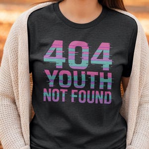 Peut inclure: T-shirt gris foncé avec l'inscription "404 YOUTH NOT FOUND" en rose, bleu et violet, avec un effet glitch. La personne porte un cardigan crème.