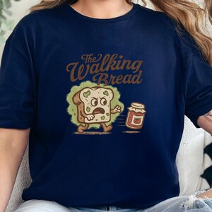 Puede incluir: Camiseta azul marino con una ilustración de dibujos animados de una rebanada de pan huyendo de un tarro de mermelada. El texto "The Walking Bread" está encima de la imagen en una fuente marrón vintage.