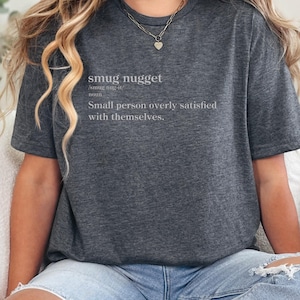Könnte beinhalten: Dunkelgraues T-Shirt mit dem Text "smug nugget" und der Definition: "Kleine Person, die mit sich selbst übermäßig zufrieden ist." Das Shirt besteht aus weichem Material.