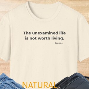 May include: A cream-coloured t-shirt featuring the quote "The unexamined life is not worth living." in dark grey text, attributed to Socrates. The word "NATURAL" is printed in yellow at the bottom of the shirt.