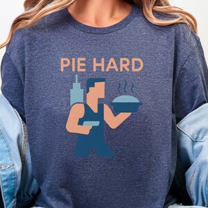 Könnte beinhalten: Marineblaues T-Shirt mit dem Text "PIE HARD" in Orange. Darunter eine Grafik einer Person, die einen Kuchen hält, mit einer Stadtlandschaft im Hintergrund. Die Grafik ist in Blau- und Orangetönen gehalten.