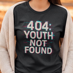 May include: Dark gray t-shirt with the text "404: YOUTH NOT FOUND" in a glitch effect. The text is white with cyan and magenta color distortions. The shirt is a crew neck style.